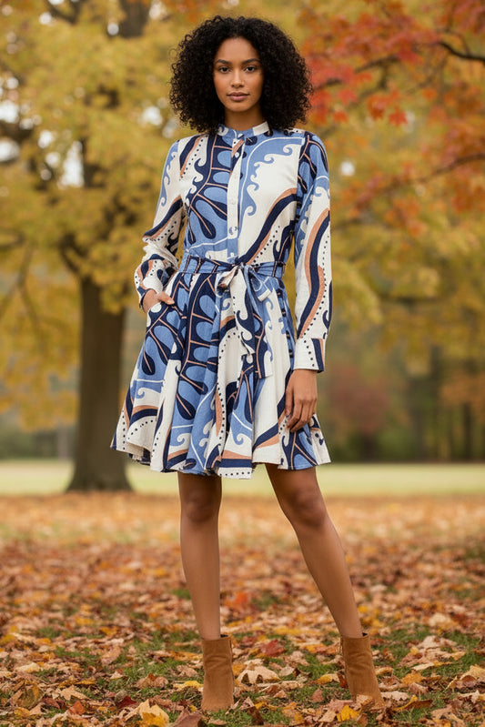 A woman modeling a short, long-sleeve dress with a blue, beige, and white abstract pattern, standing against a plain white wall.