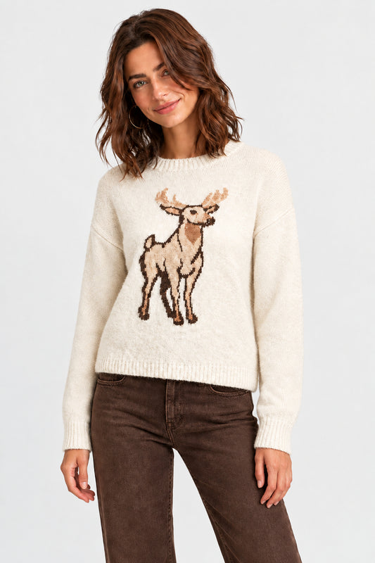 A woman wearing a cream-colored knit sweater with a detailed brown and beige embroidered deer motif on the front, paired with brown pants, against a neutral gray studio background.