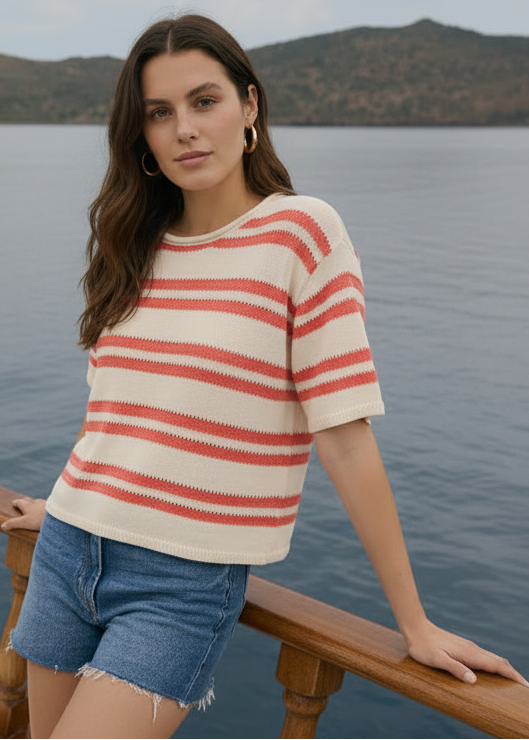 Woman wearing a red and white striped shirt and denim shorts standing on a boat with a scenic background.