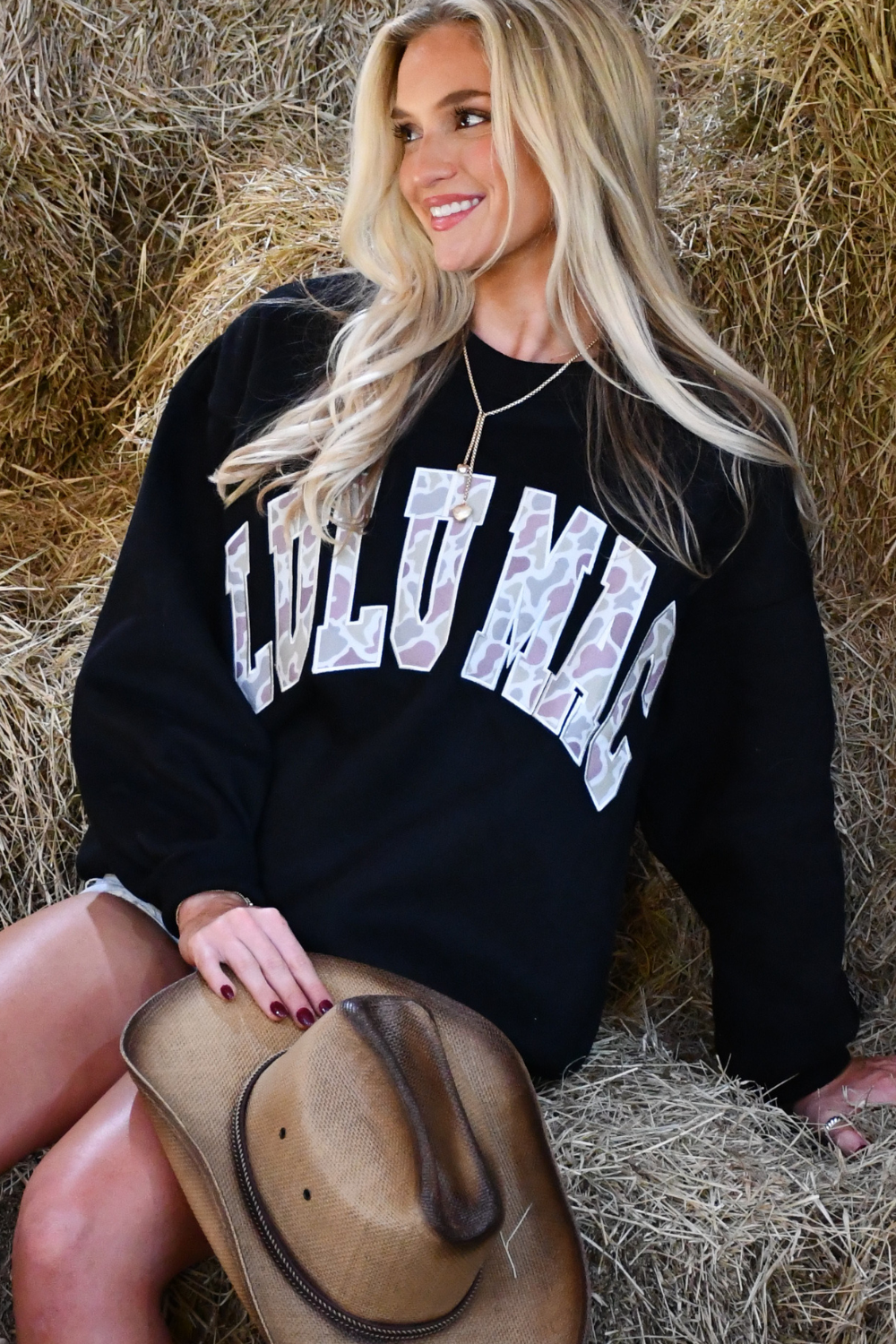Woman wearing a black sweatshirt with 'CULU MAC' text, sitting on hay.