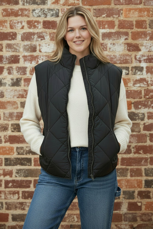 Woman wearing a black quilted vest over a white sweater and blue jeans against a brick wall.