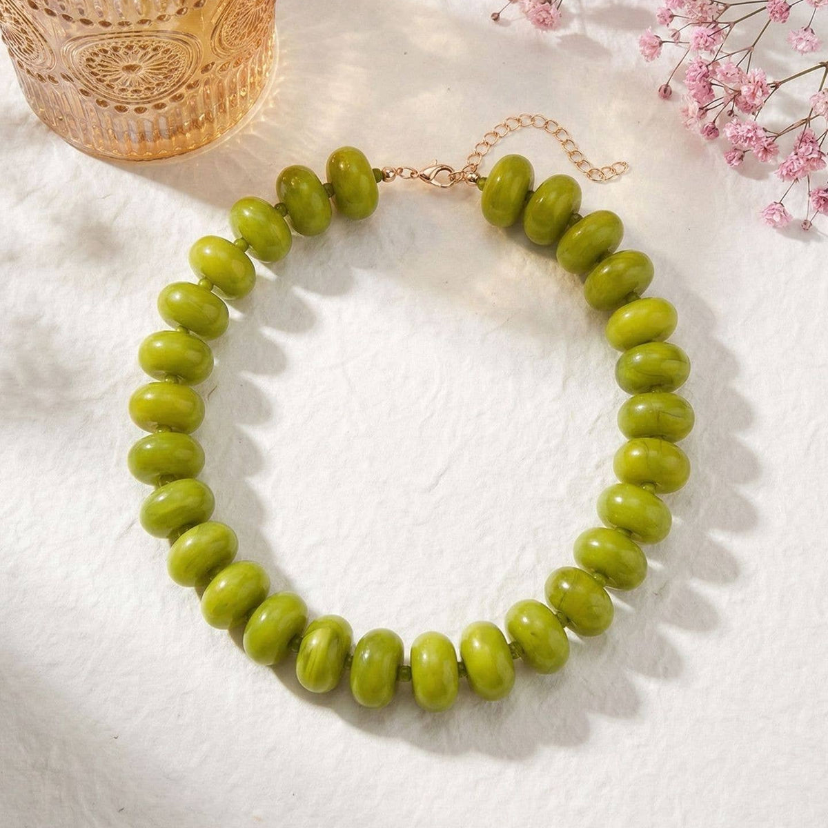 Green beaded necklace on a white surface with pink flowers and a glass container in the background.