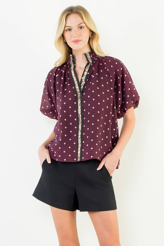 A woman modeling a short-sleeve button-up blouse in burgundy with white polka dots and a decorative gold-trimmed placket, paired with black shorts, against a plain white studio background.