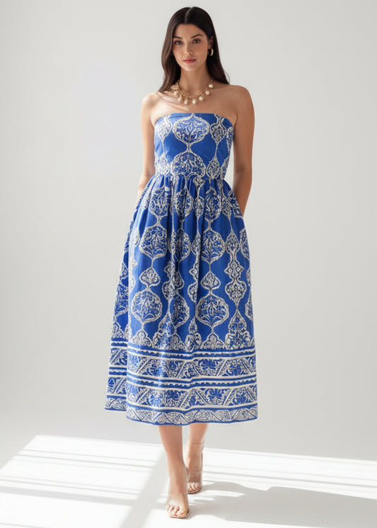 Woman wearing a blue and white patterned dress against a white brick wall.