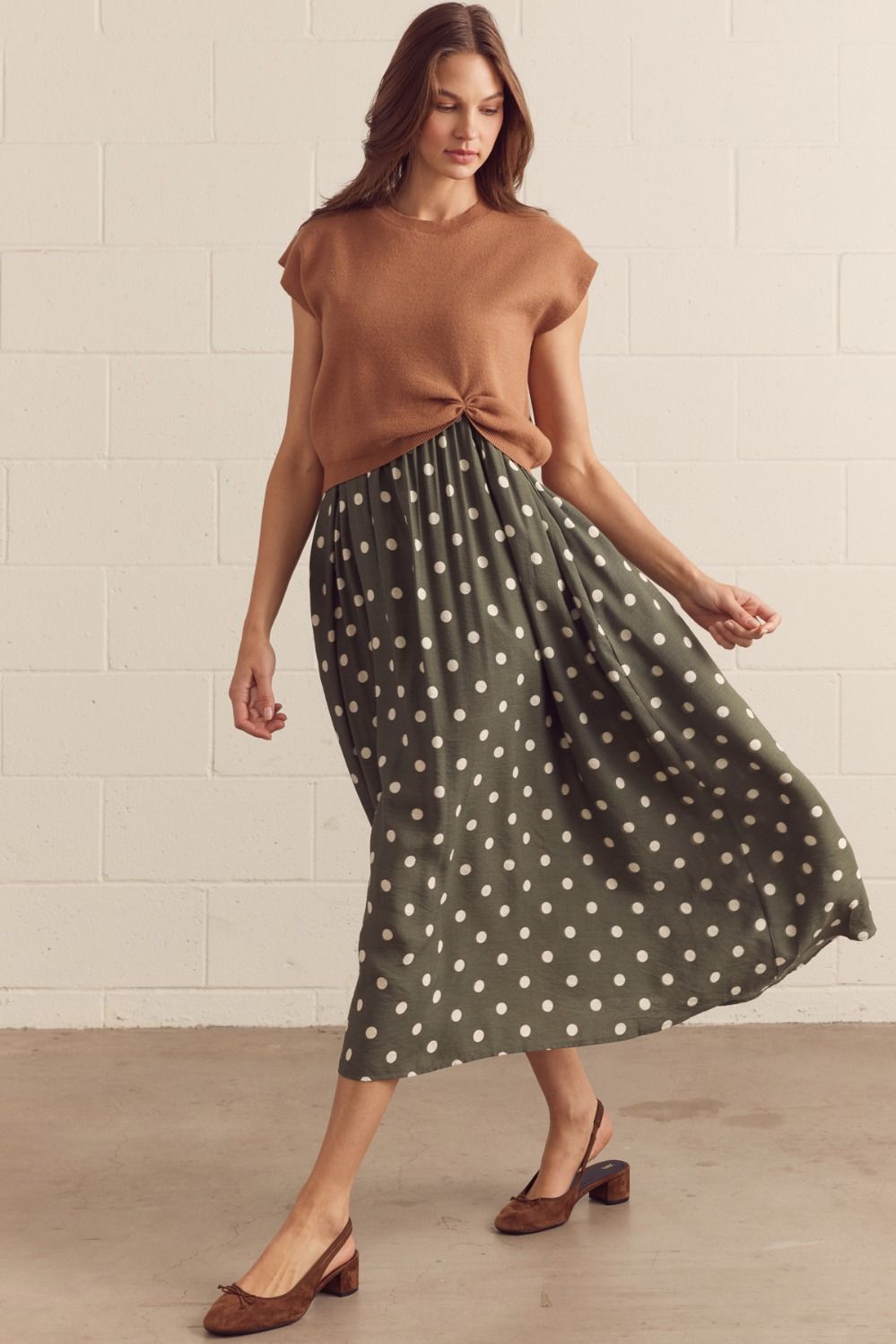 Woman wearing a brown top and green polka dot skirt against a white tiled wall.