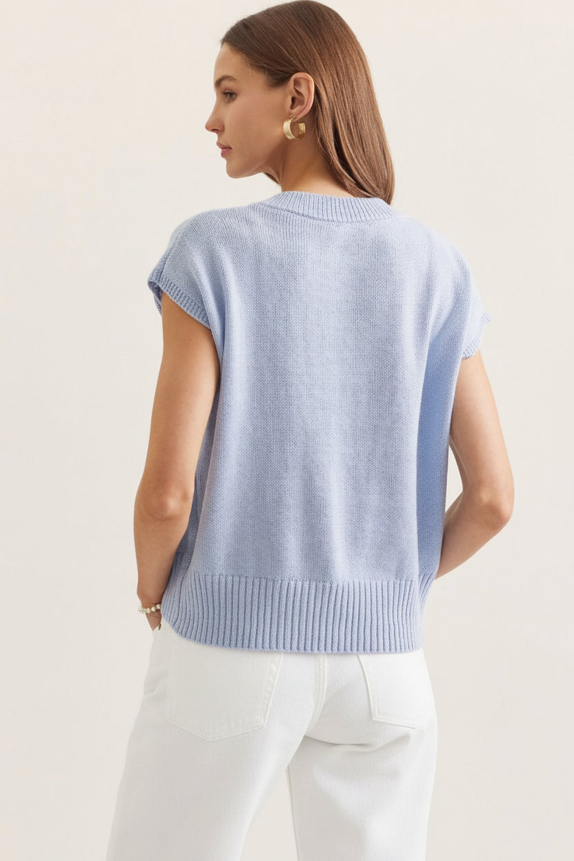 Woman wearing a light blue sleeveless sweater and gray pants on a plain background