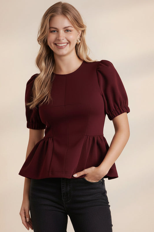 Woman wearing a burgundy peplum top and brown polka dot pants against a light brick wall.