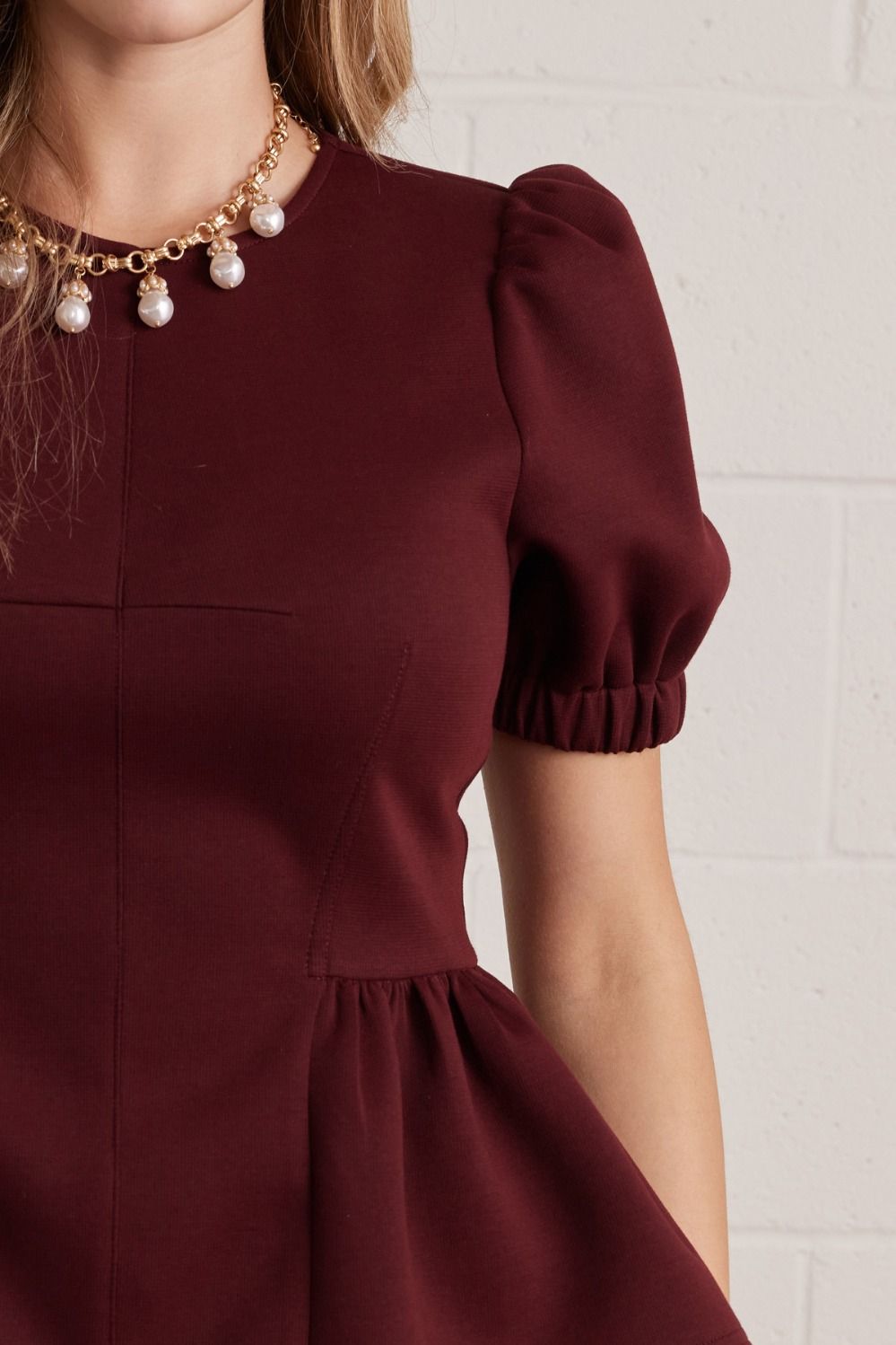 A close-up of a woman wearing a burgundy short-sleeve dress with puffed sleeves and a pearl necklace against a white brick wall.
