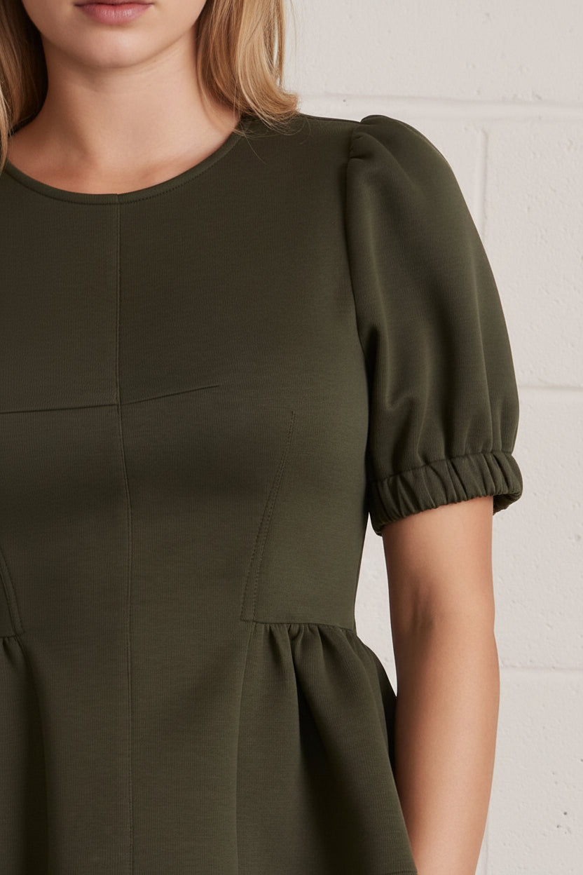 Close-up of a woman wearing an olive green peplum top with puff sleeves and a pearl necklace against a white brick wall.