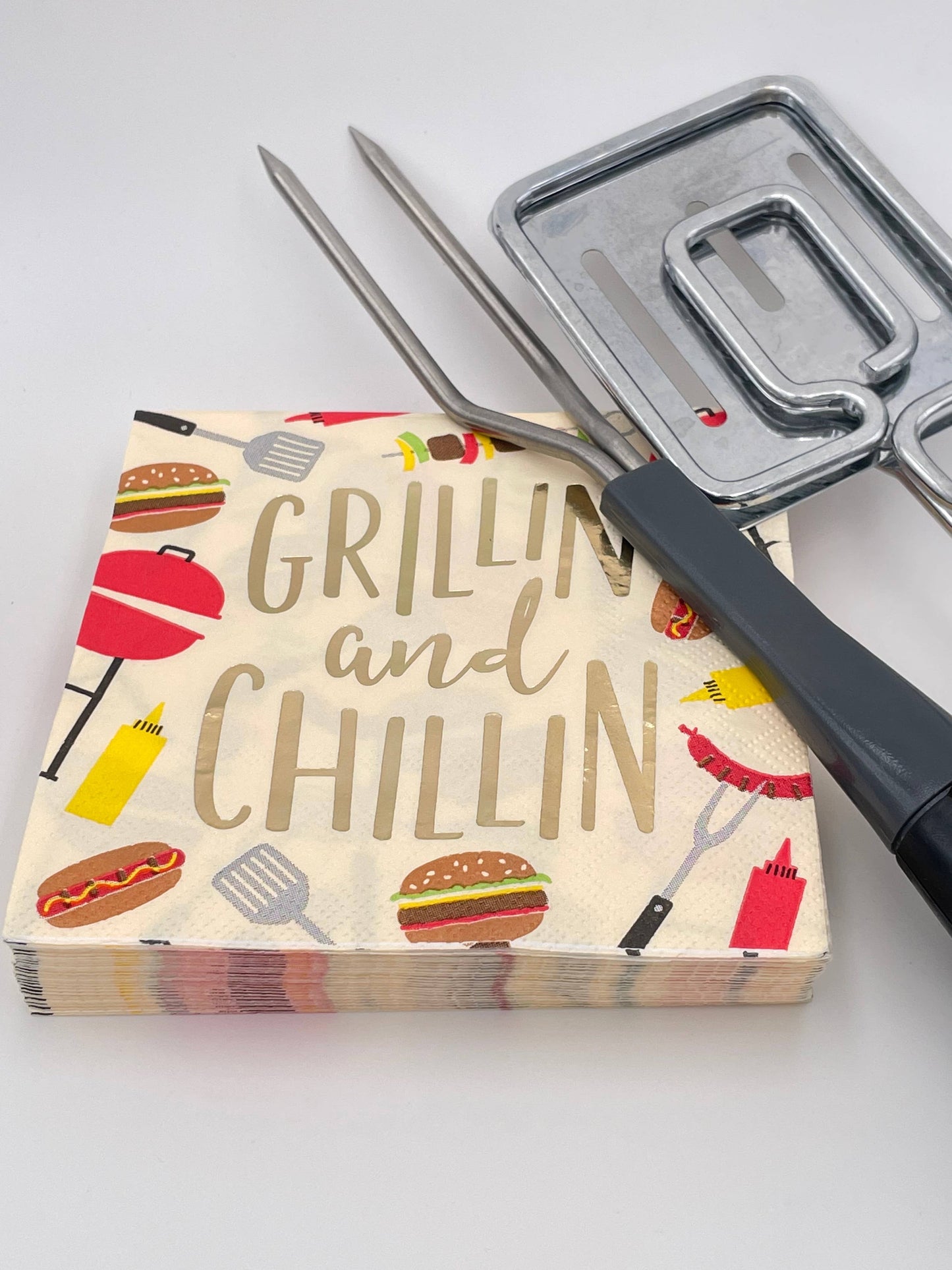 Stack of napkins with 'Grilling and Chillin' text and grill icons, next to a pair of tongs on a white background.