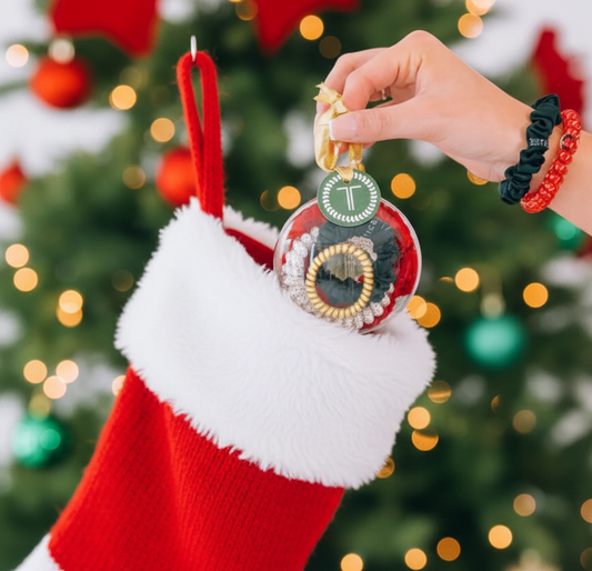 A hand holding a TELETIES holiday ornament with a Christmas tree and festive decorations in the background. The ornament is round and has text on it, and it is adorned with a red and green color scheme.